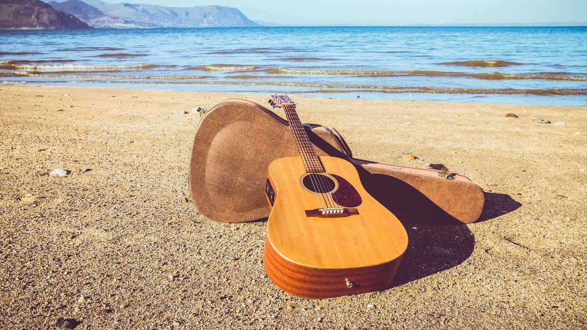 Eine Akustikgitarre liegt an einem Sandstrand, im Hintergrund das Meer und in der Ferne Klippen im blauen Dunst