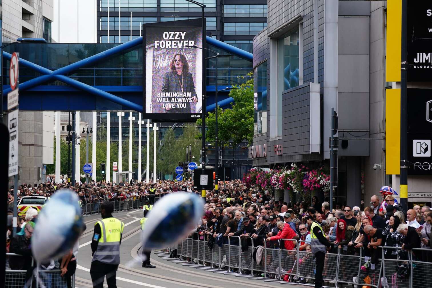 30.07.2025, Großbritannien, Birmingham: Fans warten auf der Broad Street in Gedenken an den Black-Sabbath-Frontmann Ozzy Osbourne, bevor sein Leichnam nach seinem Tod in der vergangenen Woche im Alter von 76 Jahren zu einer Prozession in seine Heimatstadt zurückgebracht wird. Foto: Jacob King/PA Wire/dpa +++ dpa-Bildfunk +++