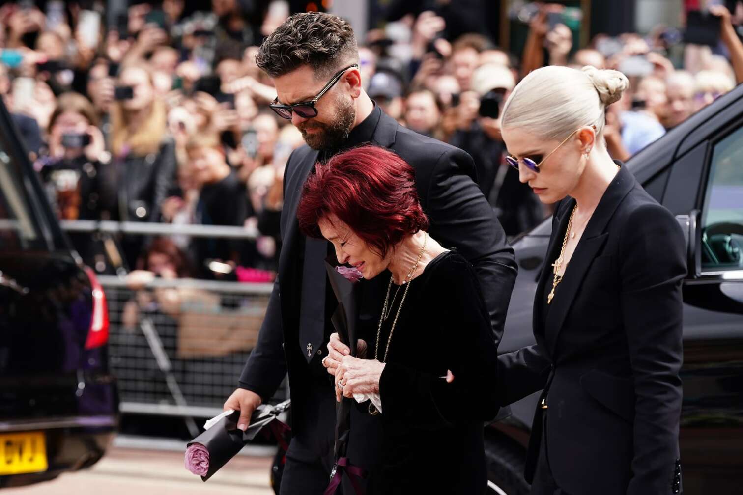 30.07.2025, Großbritannien, Birmingham: Die Familie von Ozzy Osbourne, Jack Osbourne (l-r), Sharon Osbourne und Kelly Osbourne legen Blumen nieder und betrachten die Botschaften und Blumenspenden, die an der Black-Sabbath-Brücke in der Broad Street zum Gedenken an den Black-Sabbath-Frontmann Ozzy Osbourne hinterlassen wurden, während sein Leichnam nach seinem Tod letzte Woche im Alter von 76 Jahren für eine Prozession in seine Heimatstadt zurückgebracht wird. Foto: Jacob King/PA Wire/dpa +++ dpa-Bildfunk +++