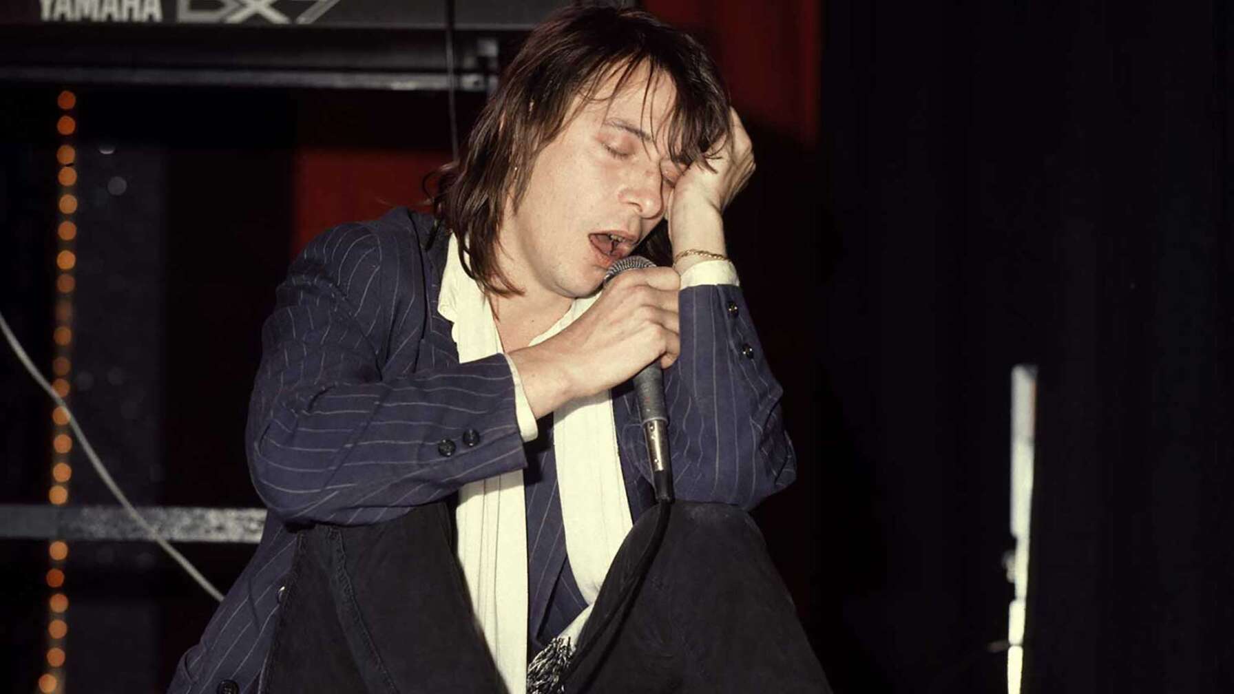 Ton Steine Scherben-Sänger Rio Reiser sitzt mit einem Mikrofon in der Hand vor einem Gitarrenverstärker.